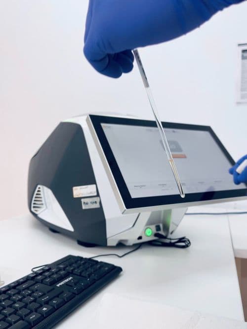 A gloved hand holding a testing tool over a touch-screen laboratory analysis machine, with a keyboard in the foreground.